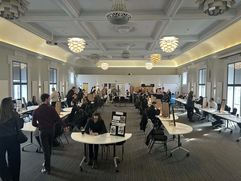 Tables around a busy room with participants sitting at each table with a notice board on it.